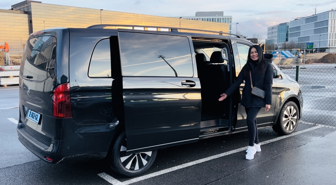 A taxi was parked in the parking lot. The door opened. A woman stood beside the door, smiling, extending her hand to invite passengers to get in the car.